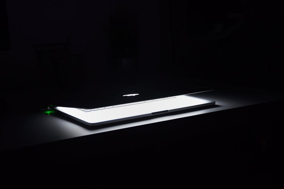 A dramatic, low-light image of a slightly open laptop with glowing keys on a dark desk